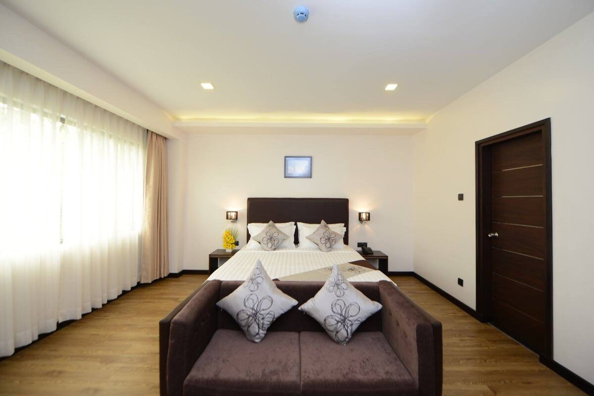 Ein Schlafzimmer mit einem großen Bett, zwei Nachttischen, einer Kopfteil, einem Fenster mit Vorhängen, einer Tür und einem braunen Sofa mit Zierkissen.