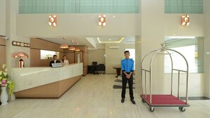 A hotel reception area with a reception desk, two receptionists, a man standing, and a cart. - Hotel Lexus Yangon (Yangon)