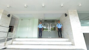 Two uniformed individuals standing on a marble staircase at the entrance of a building with glass doors and a security camera above. - Hotel Lexus Yangon (Yangon)