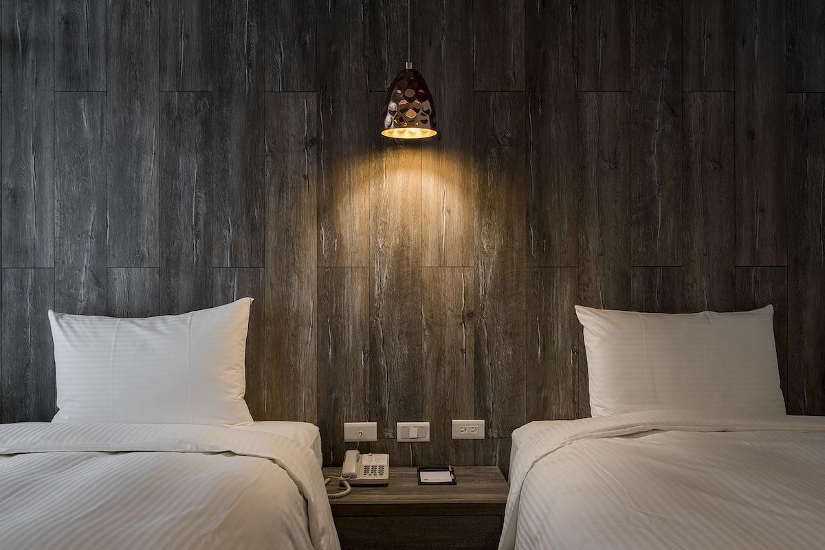 Quarto de hotel com duas camas, uma mesa de cabeceira com um telefone e um tablet, e uma luminária fixada na parede.
