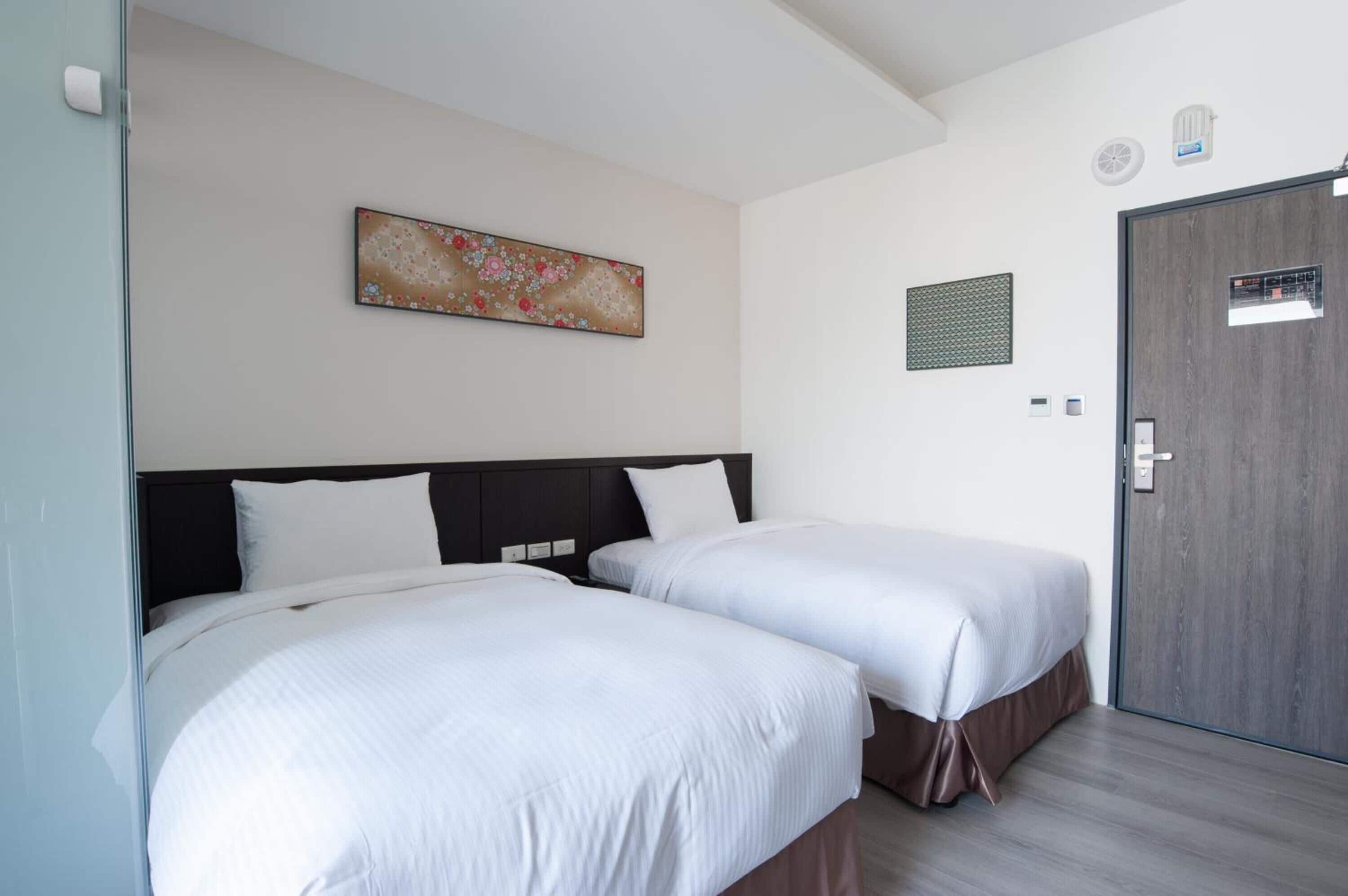 A hotel room with two beds, a wooden door, a wall-mounted air conditioning unit, and a floral artwork.