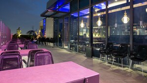 Un espacio al aire libre con mesas y sillas, ventanas amplias e iluminación decorativa.