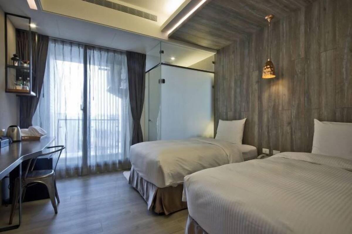 Quarto de hotel com duas camas, uma escrivaninha e uma janela grande com cortinas.