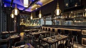 Un restaurante moderno con varias mesas y sillas, iluminación colgante y una zona de bar con botellas y vasos.