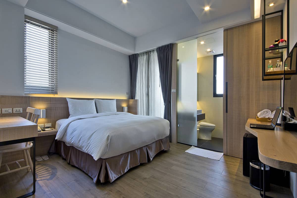Quarto de hotel moderno com uma cama grande, uma escrivaninha, um laptop e um banheiro visível através de uma porta de correr.