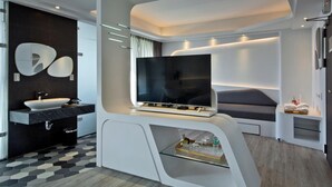 Habitación de hotel moderna con un televisor de pantalla plana grande, una consola blanca con repisa de vidrio, un baño blanco y negro y un sofá gris.