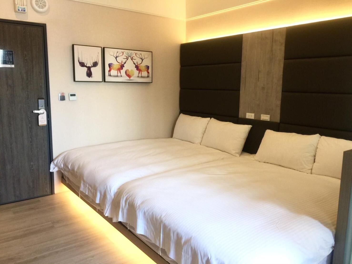 Un lit bien fait, avec une tête de lit sombre et du linge de lit blanc, une porte dotée d’une serrure, et deux œuvres d’art représentant des cerfs, encadrées et accrochées au mur.
