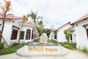 A bungalow with a tiled roof, palm trees, and a central stone feature with the text "WINGS Bungalow". - Wings Bungalow (Phu Quoc)