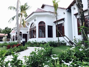 A white building with a tiled roof, arched windows, and a small garden area with palm trees and flowering plants. - Wings Bungalow (Phu Quoc)