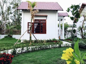 A white building with a red-tiled roof, a window with a wooden shutter, and a small garden with red flowers and a yellow plant. - Wings Bungalow (Phu Quoc)