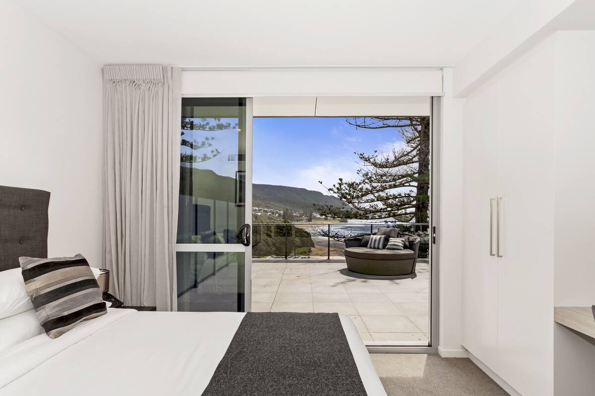 A modern bedroom with a large bed, a glass door leading to a balcony with a view of a mountain and a body of water.