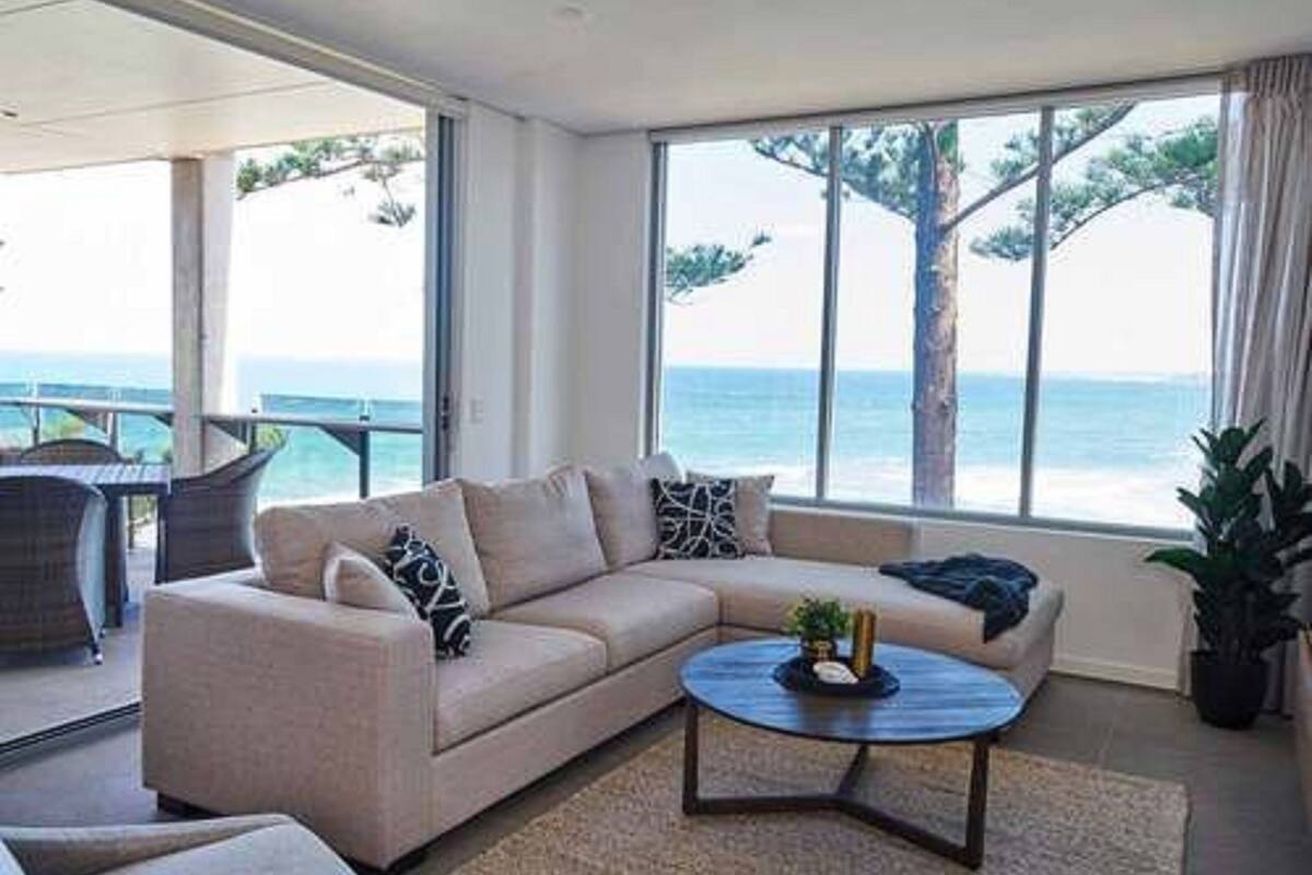 A modern living room with a beige sofa, a round coffee table, and a view of the sea through large windows.