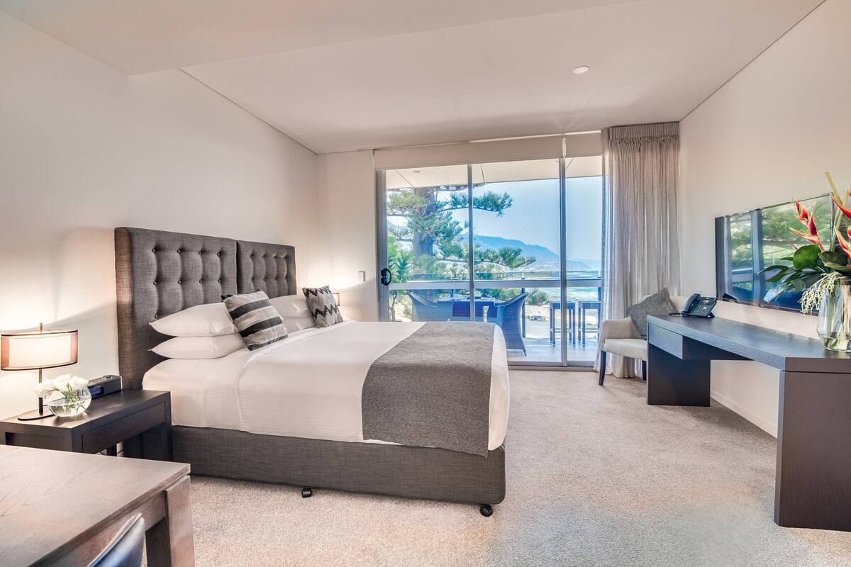 A modern hotel room with a large bed, a desk, a chair, and a balcony with a view of the sea.