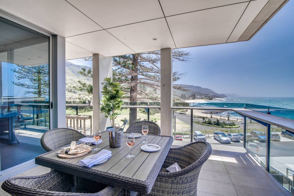 A balcony with a table set for two, overlooking a coastal view with mountains in the background.