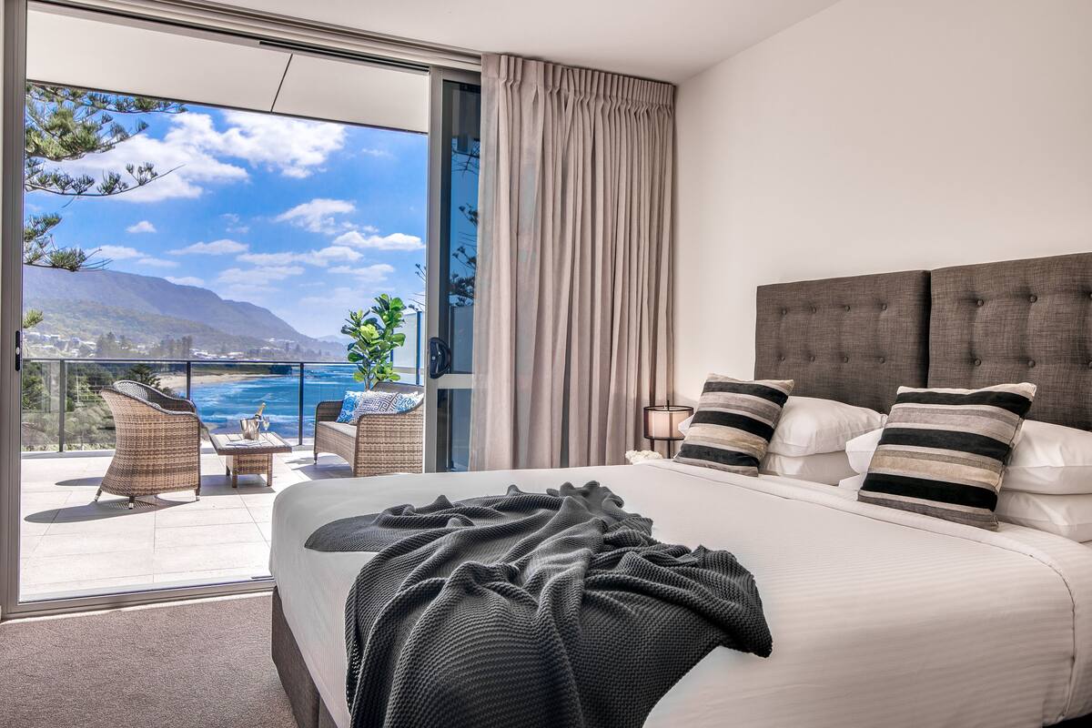 A bedroom with a large bed, a view of the ocean, and a balcony with wicker furniture.