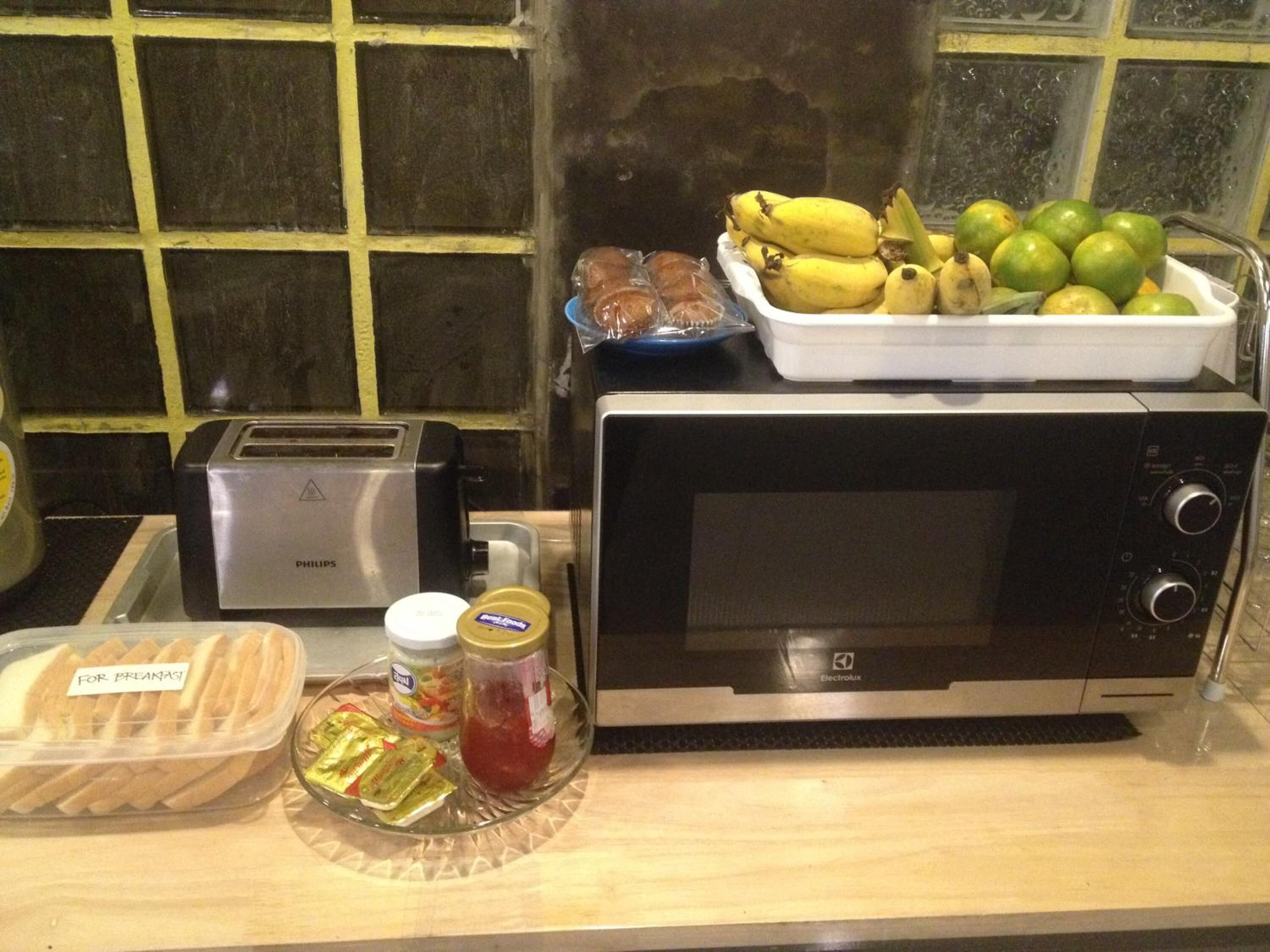 A kitchen counter with a toaster, a microwave, a tray of bread, a jar of jam, and a bowl of fruit.