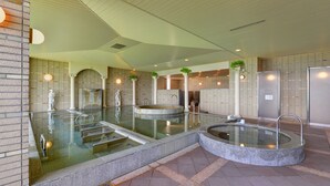 A modern indoor pool area with multiple water features, including a central pool and smaller hot tubs, surrounded by stone walls and columns. - Hotel Azalea (Tsu)