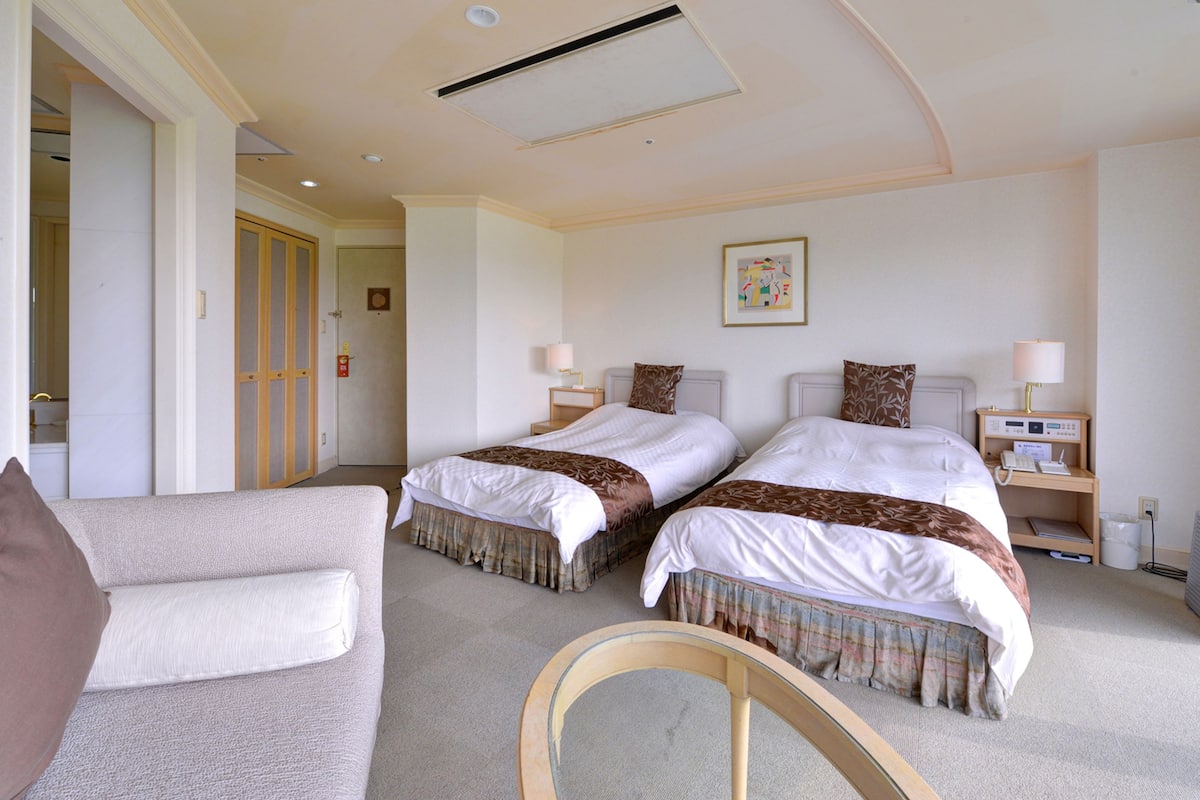 A hotel room with two beds, a sitting area with a chair, and a small desk with a lamp and phone.