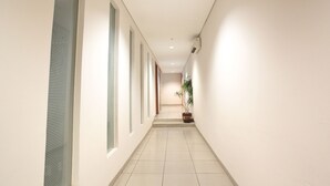 A long, narrow hallway with white walls and a tiled floor. There are several windows along the left wall and a potted plant in the center. - LeGreen Suite Kuningan (Jakarta)