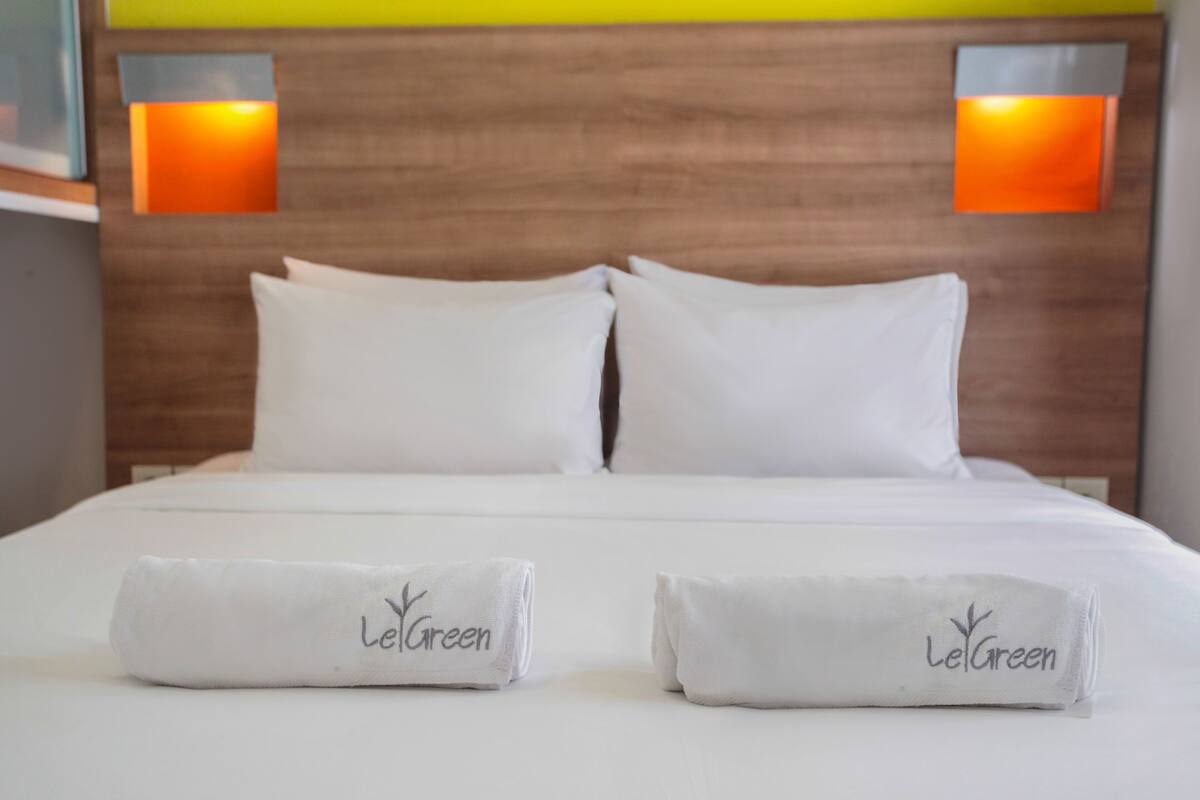 A neatly made bed with two pillows and two rolled towels with 'LetGreen' embroidered on them, set against a wooden headboard with two wall-mounted lights.