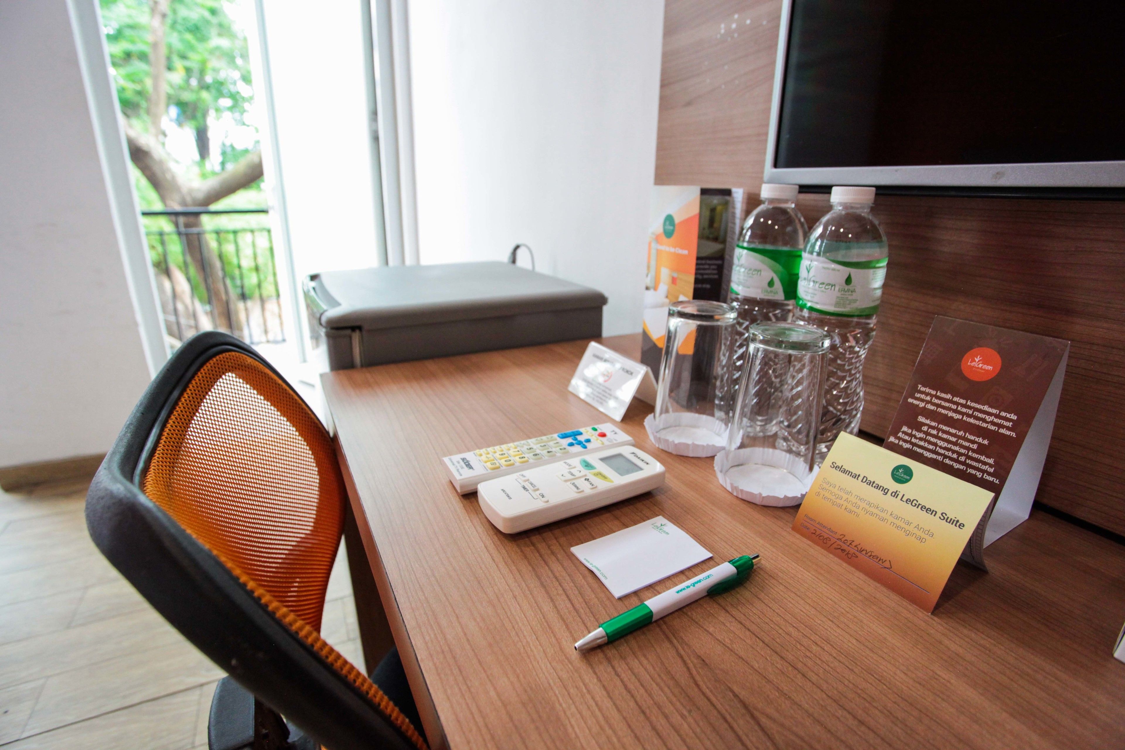 A hotel room with a wooden desk, a chair, a calculator, a pen, a glass of water, and a sign.