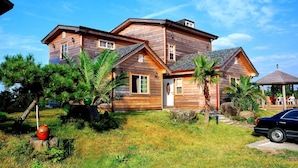 Une maison en bois à deux étages, dotée d’un porche à l’avant, entourée d’une cour verdoyante et de palmiers.