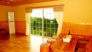 Un intérieur en bois avec une grande fenêtre, un réfrigérateur, un banc en bois et une table sur laquelle se trouvent des fruits et une bouteille.