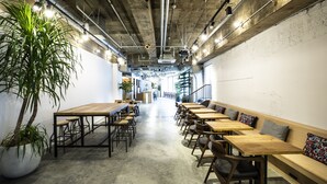 A modern interior with exposed concrete ceilings, wooden tables and benches, and a large potted plant. - HafH Fukuoka THE LIFE - Hostel (Fukuoka)