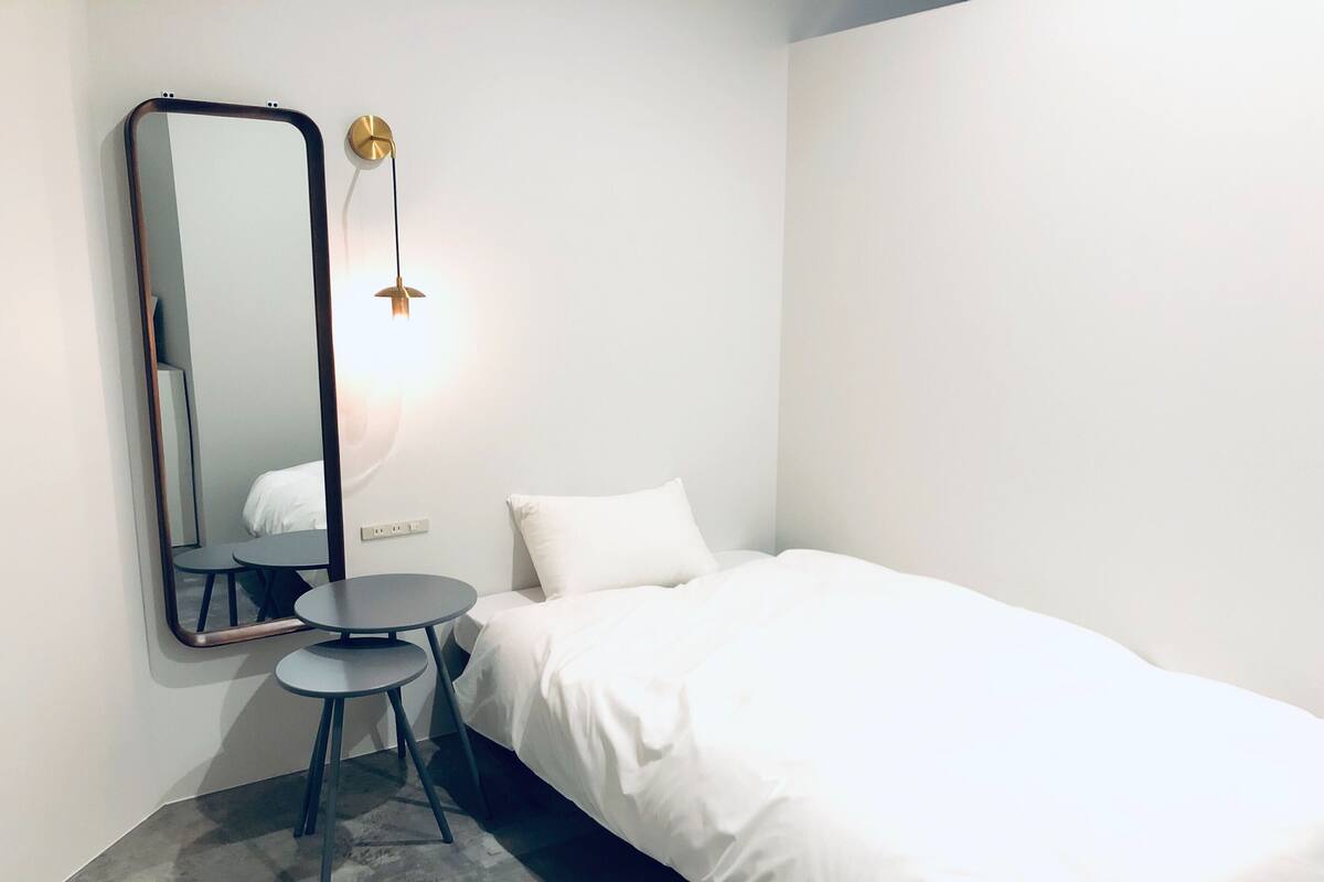 A minimalist hotel room with a single bed, a small round table, a wall-mounted mirror, and a simple light fixture.