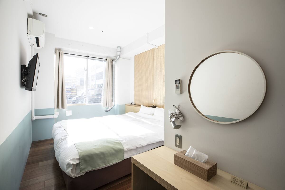 A hotel room with a bed, a bedside table with a tissue box, a mirror, and a window with curtains.