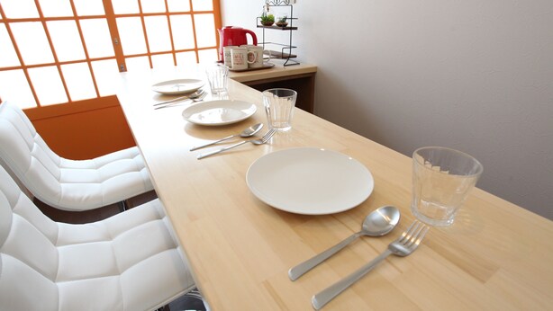 Una mesa de comedor dispuesta con platos, cubiertos y vasos, con una tetera roja y una planta pequeña en un estante al fondo.