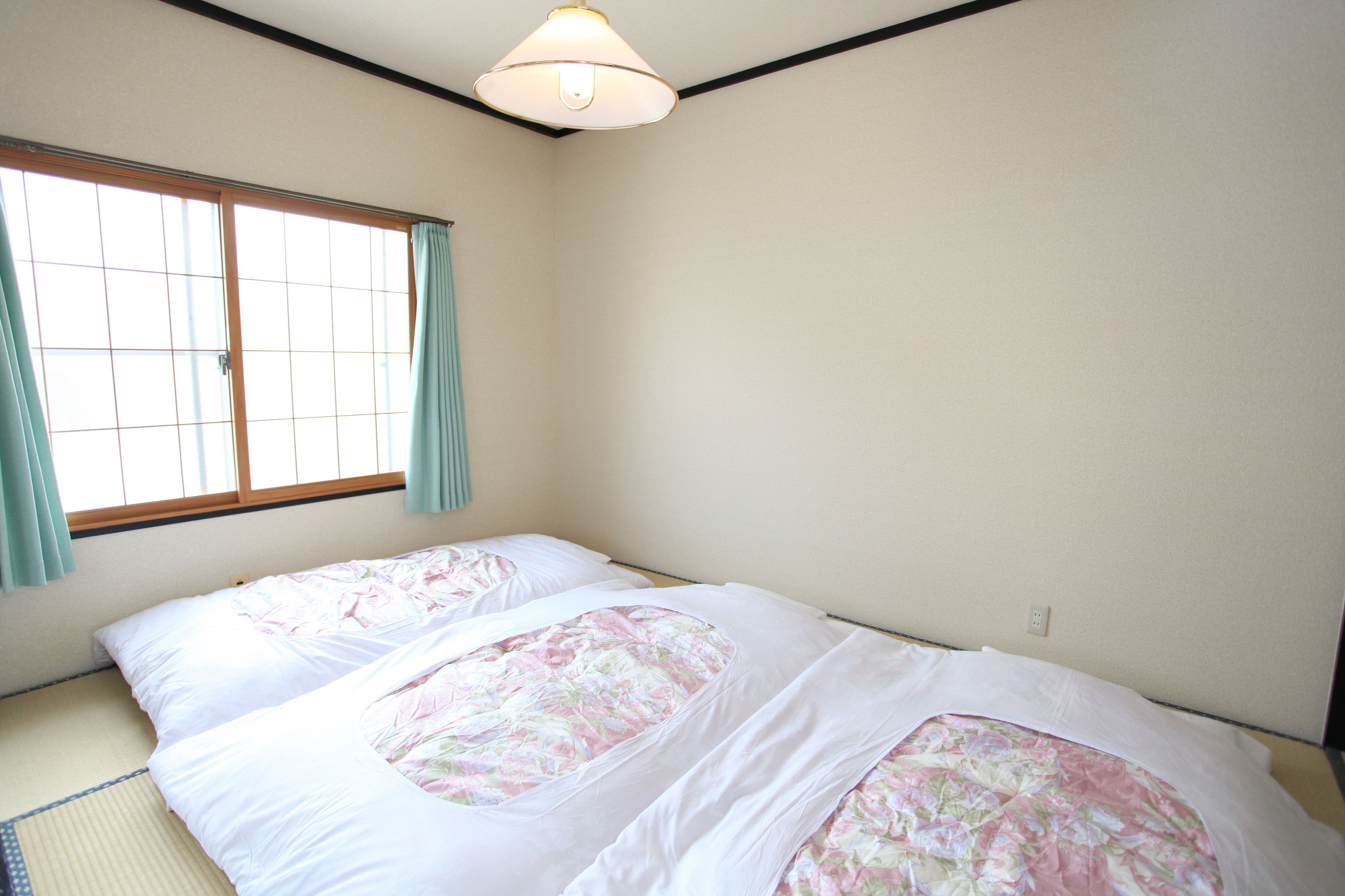 A room with tatami flooring, a bed with floral bedding, a window with curtains, and a ceiling light fixture.