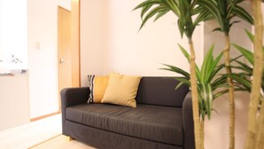 Un sofá oscuro con un cojín amarillo, una puerta de madera y una planta en maceta en una habitación con paredes de colores claros y suelo de madera.