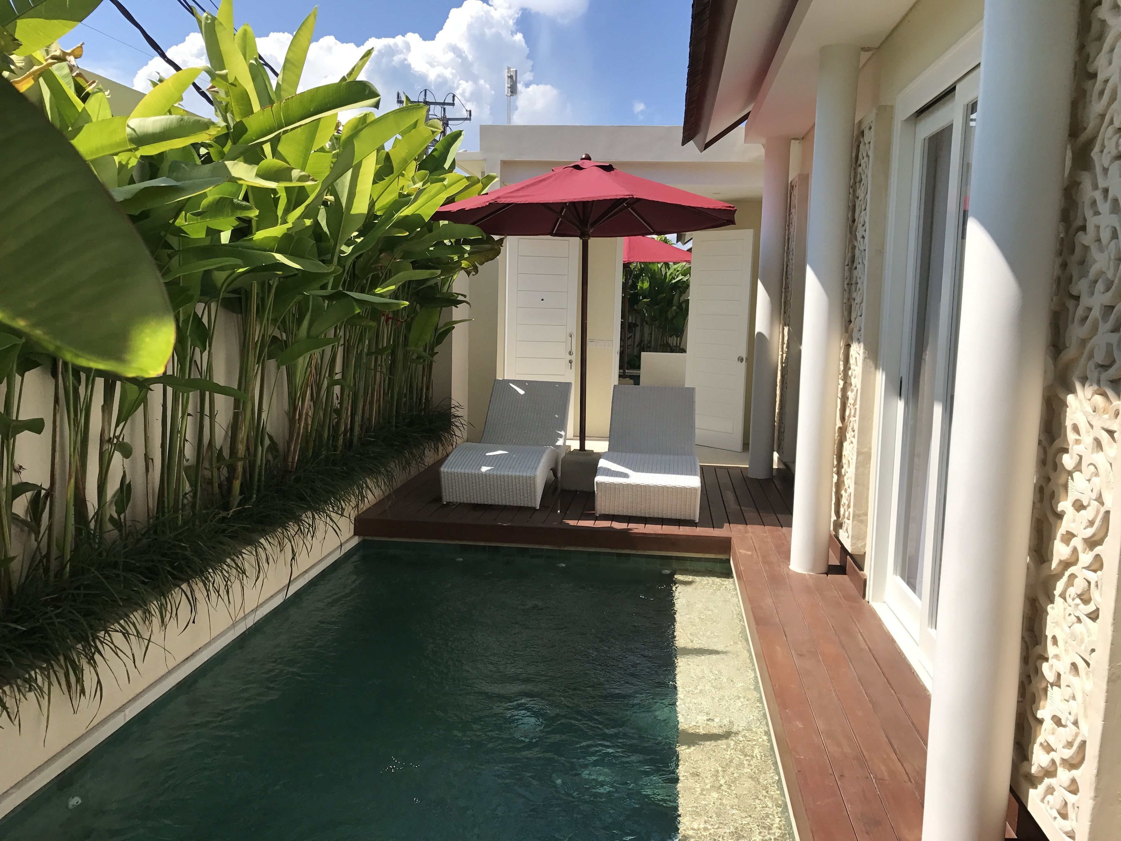 A pool area with two lounge chairs and a red umbrella, surrounded by lush greenery and white walls.