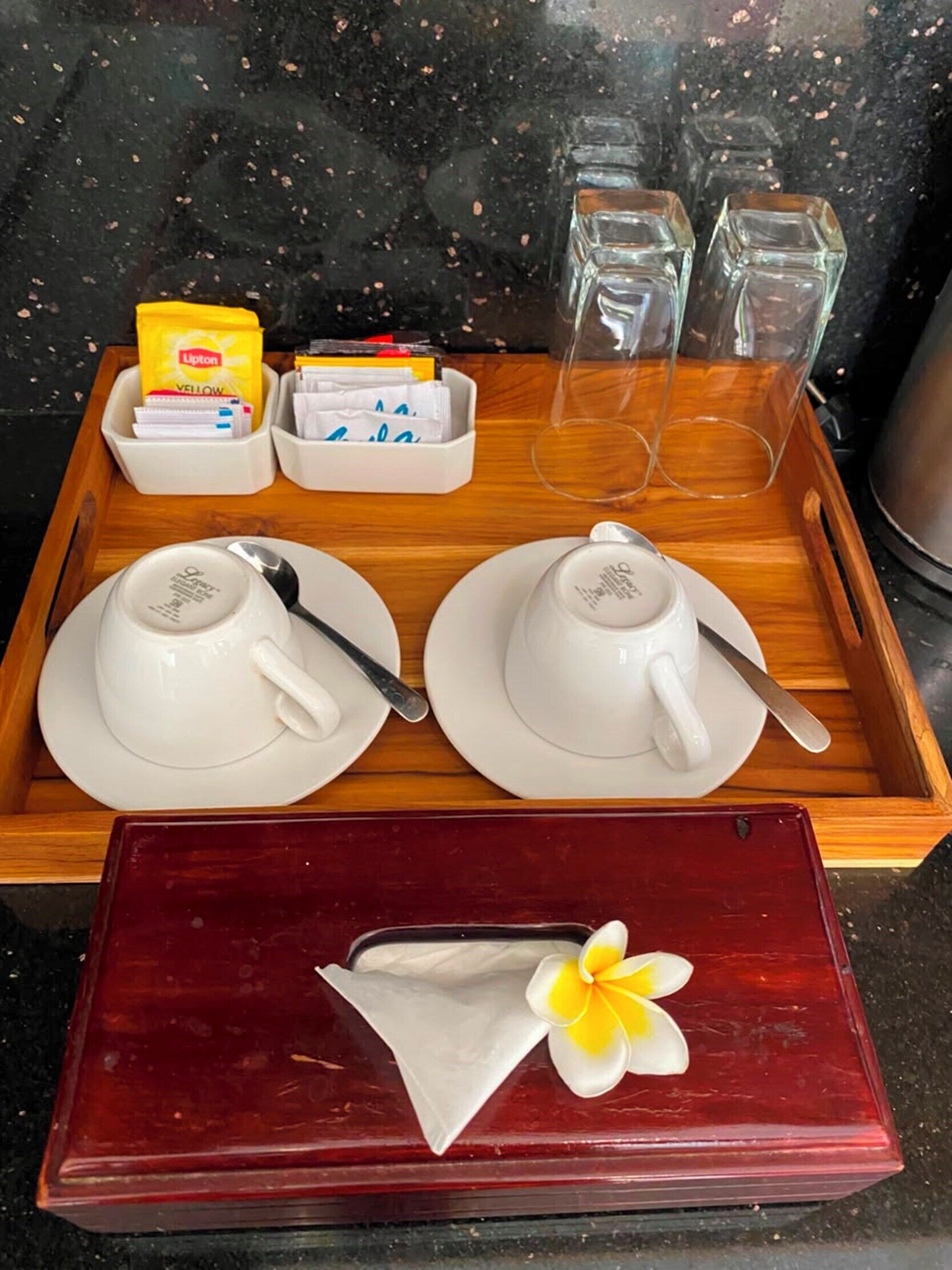 Un plateau avec deux théières blanches, deux cuillères, un paquet jaune et une boîte rouge contenant une serviette blanche et une fleur jaune.