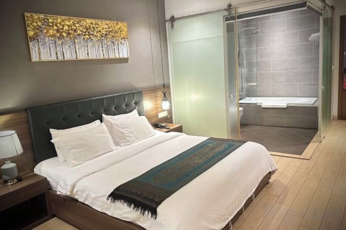 Habitación de hotel con cama, mesita de noche, lámpara, zona de ducha y puerta de cristal que da al baño.