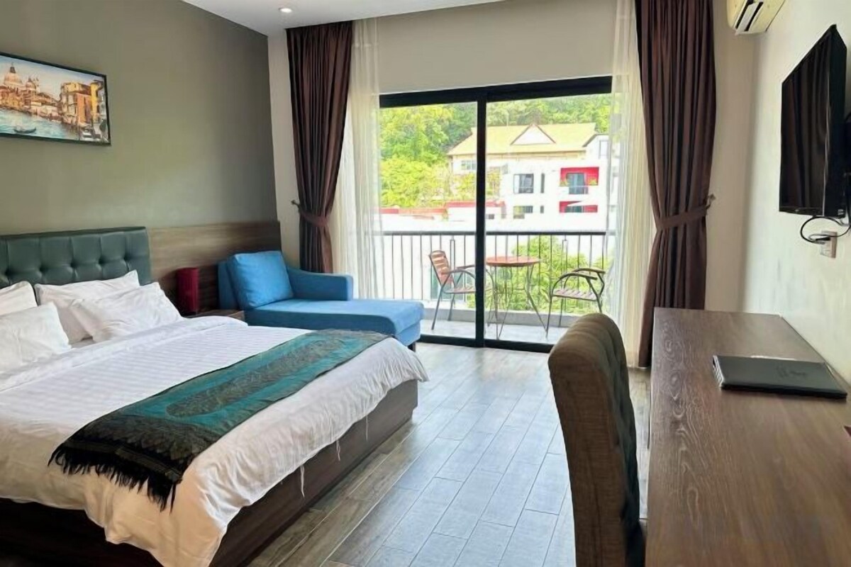 A hotel room with a bed, a desk, a chair, a balcony with a view, and a painting on the wall.