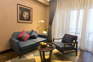 A living room with a blue sofa, a wooden chair, a small round table with a fruit bowl, and a framed picture on the wall.
