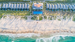 Vista aérea de un resort con una gran piscina, villas frente a la playa y una playa de arena.