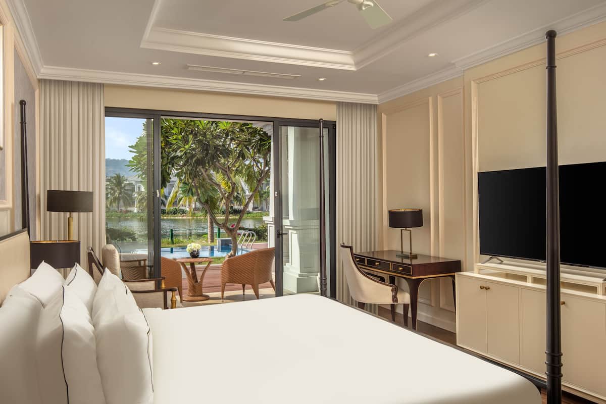 A hotel room with a bed, a desk, a television, and a view of a pool and greenery.
