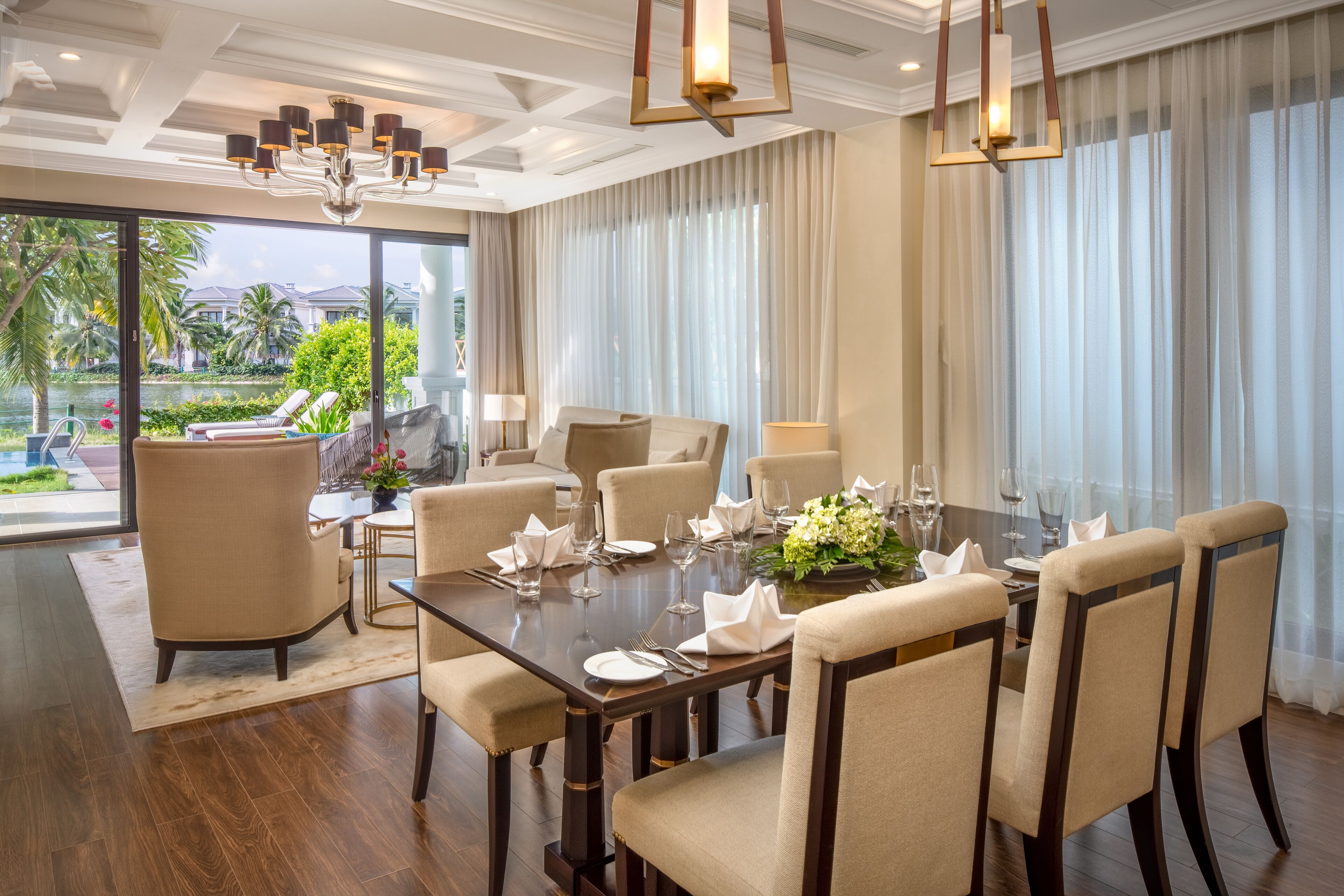 A dining area with a large table set for six, surrounded by six chairs, with a view of a pool and palm trees outside.