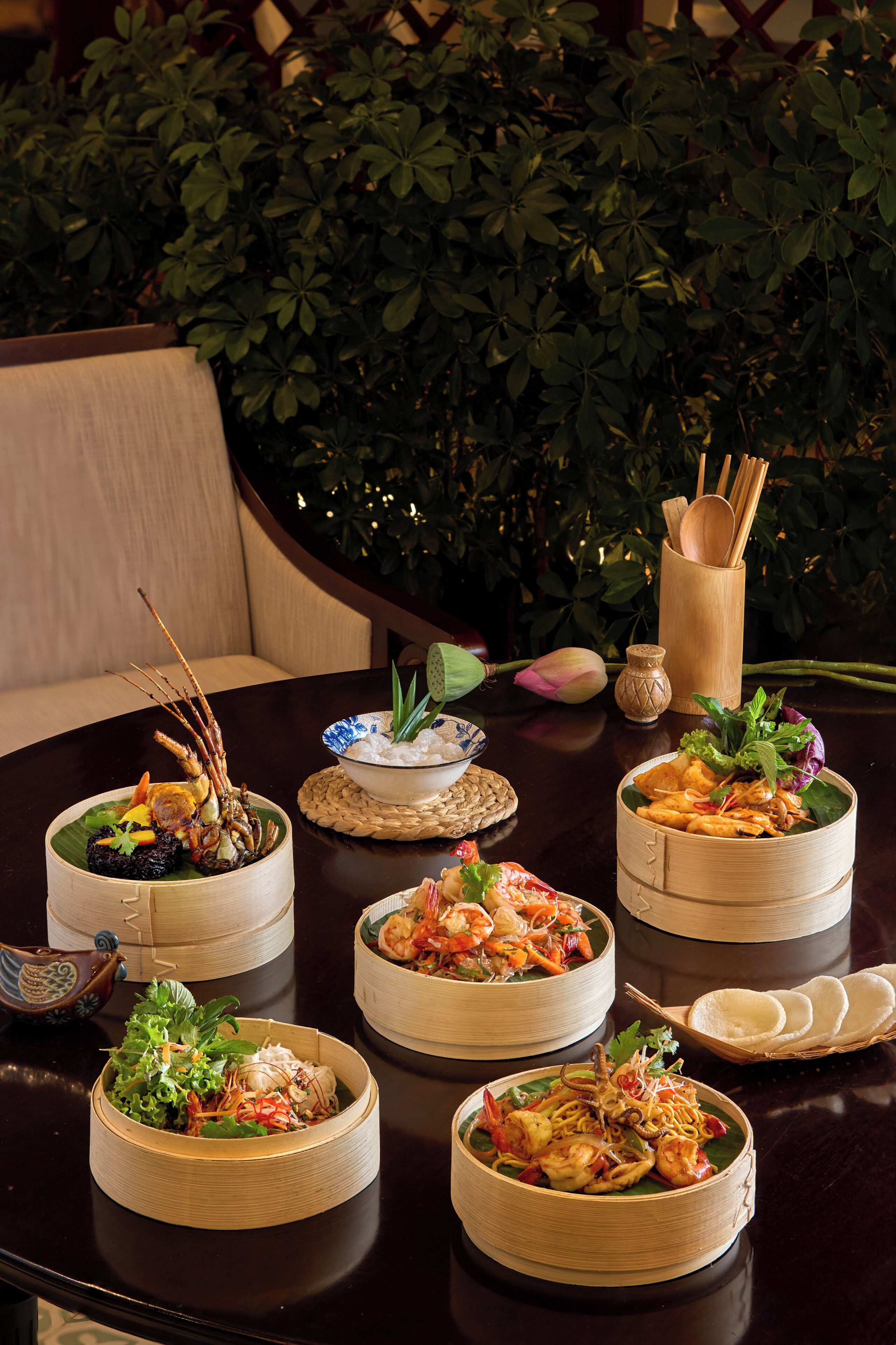 A table with various dishes in bamboo containers, a woven placemat, and a wooden spoon holder.