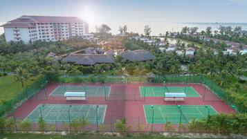 Et tenniskompleks med flere baner, omgivet af frodig vegetation og et stort hotel i baggrunden.