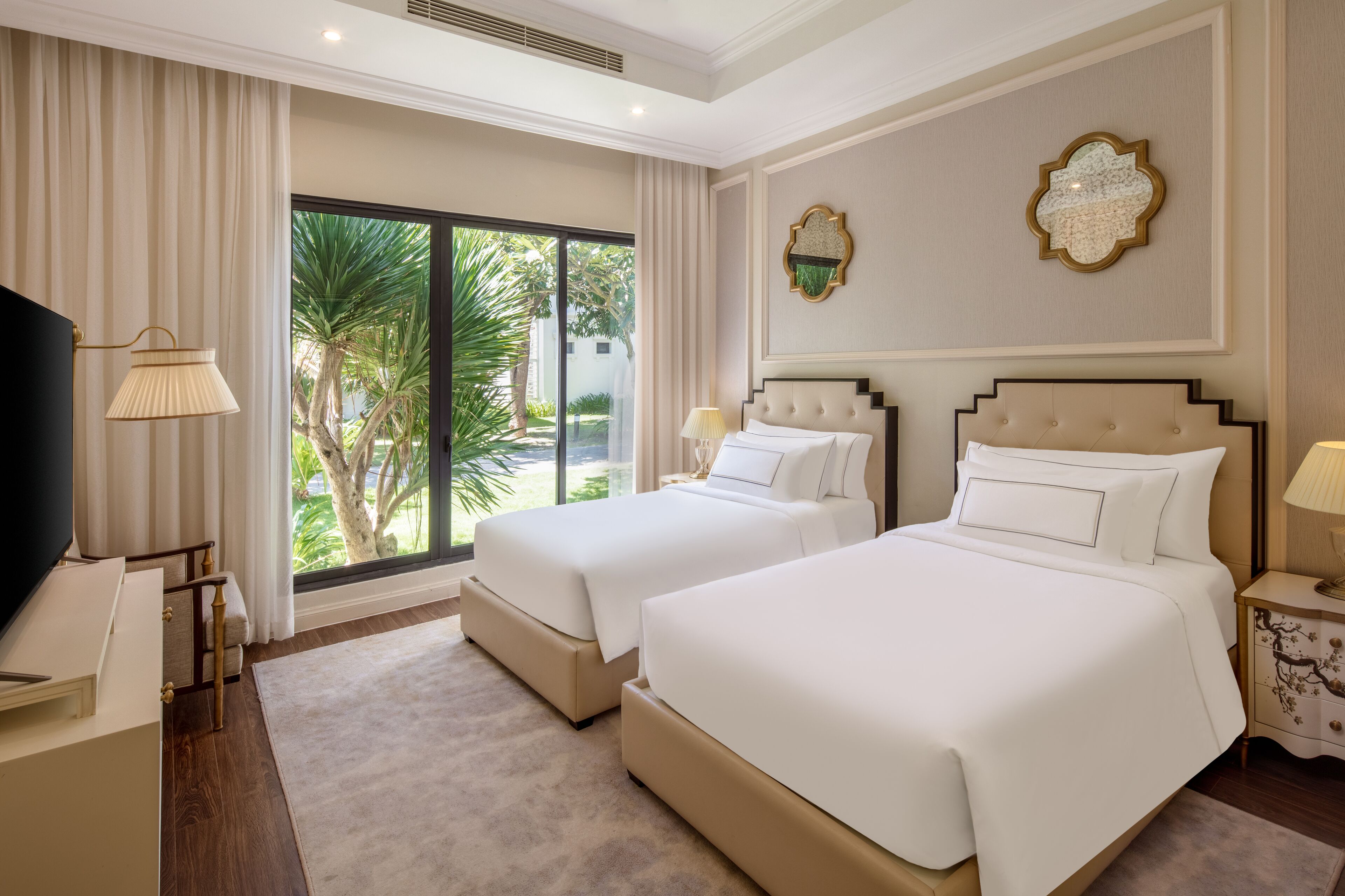 A hotel room with two beds, a TV, a desk, and a large window with a view of palm trees.