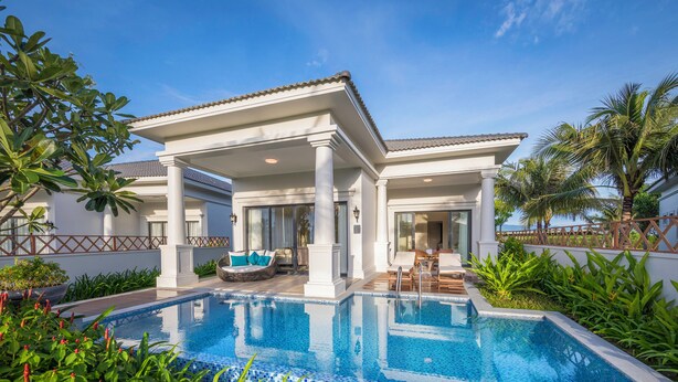 Un área junto a la piscina con una piscina azul cristalina, columnas blancas y un espacio de descanso cubierto.