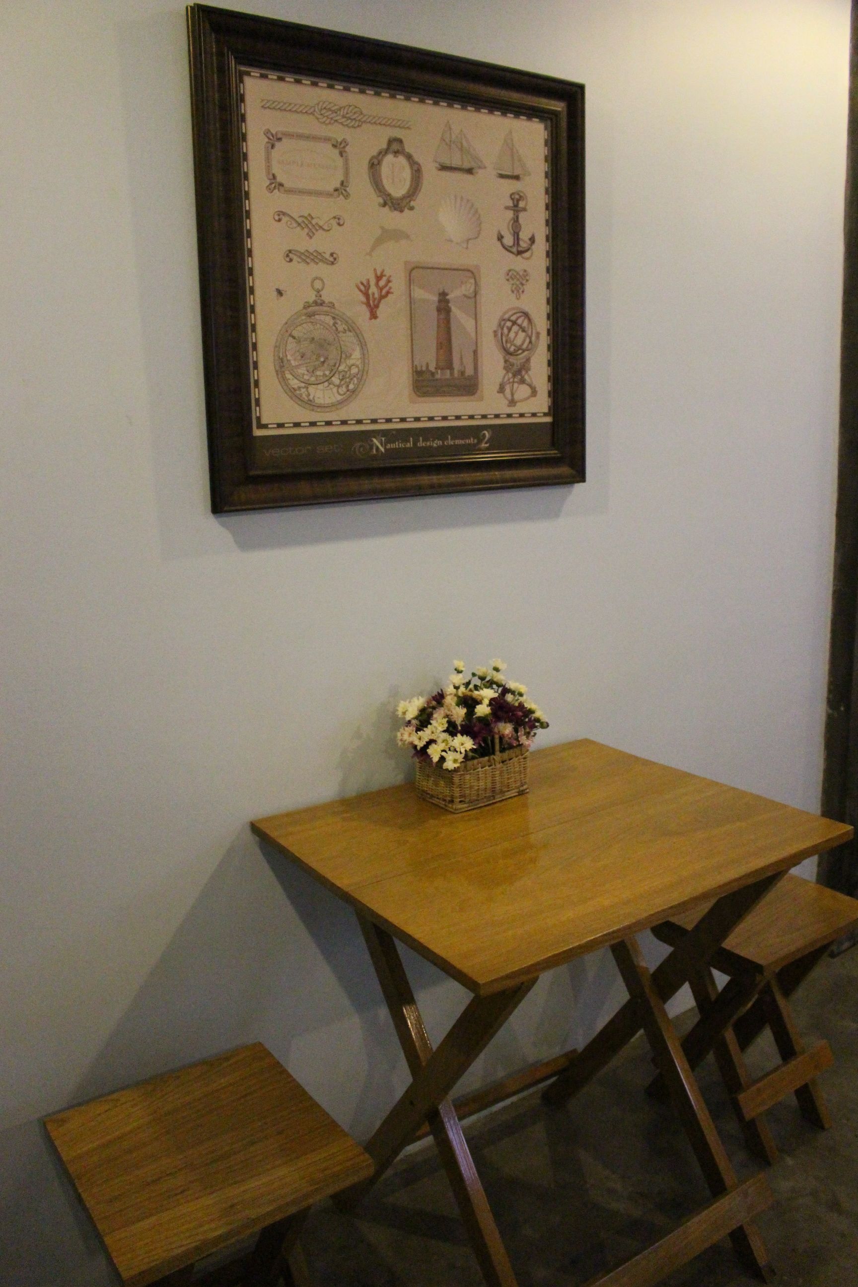 A framed nautical-themed artwork on a wall, a wooden table with a basket of flowers, and a small wooden stool.