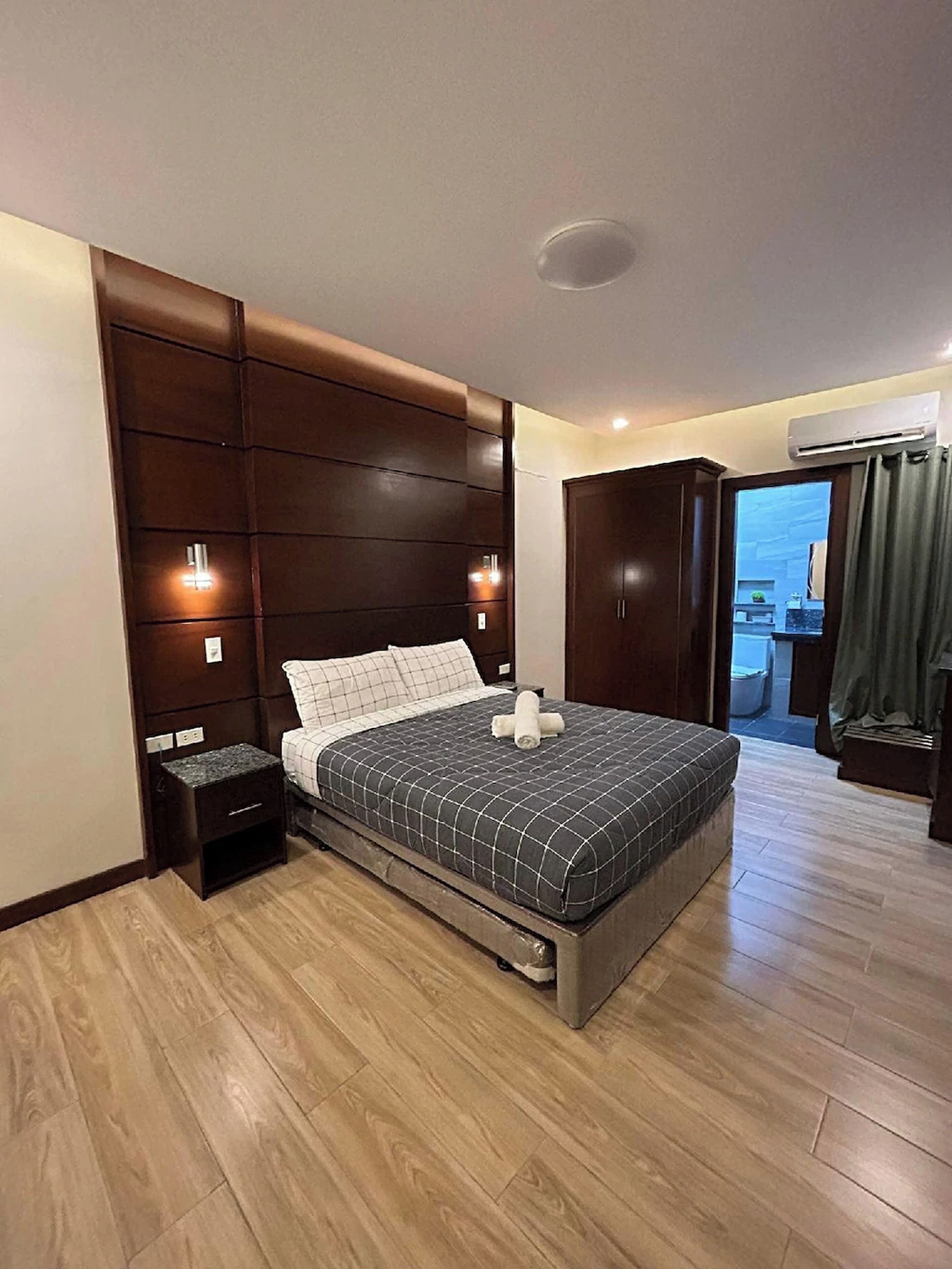 A modern bedroom with a large bed, wooden headboard, bedside tables, and an air conditioning unit.