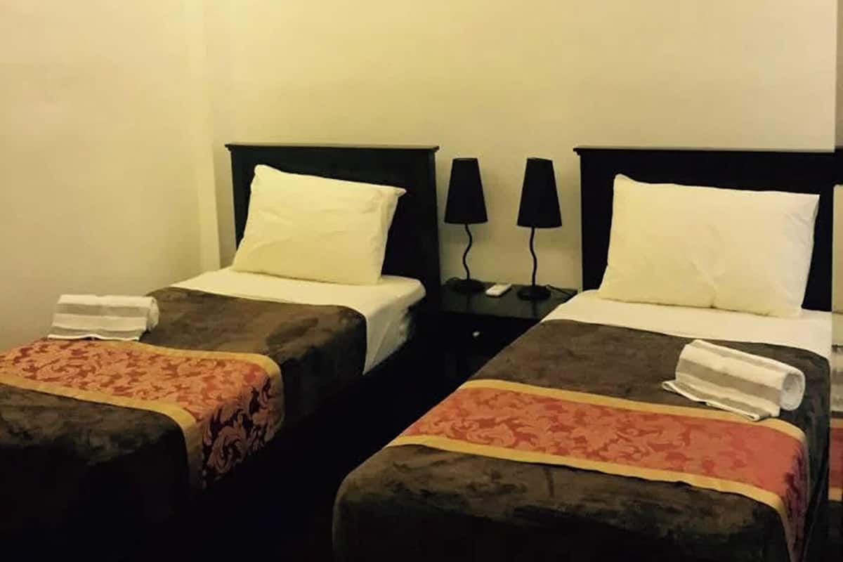 A hotel room with two single beds, each with a pillow and a blanket. There is a nightstand with a lamp between the beds.