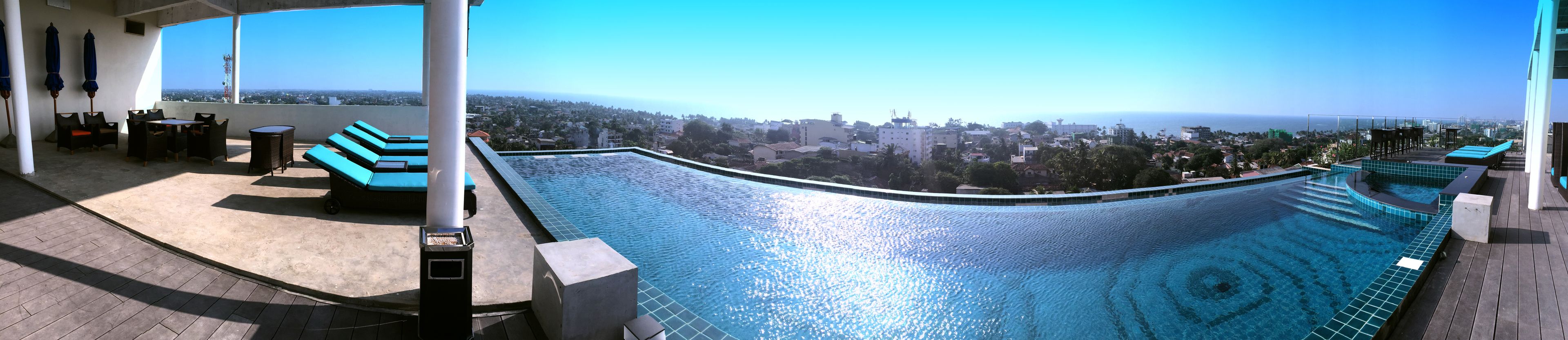 Una piscina en la azotea con borde curvo, un área de descanso con tumbonas azules y vistas a un paisaje urbano.