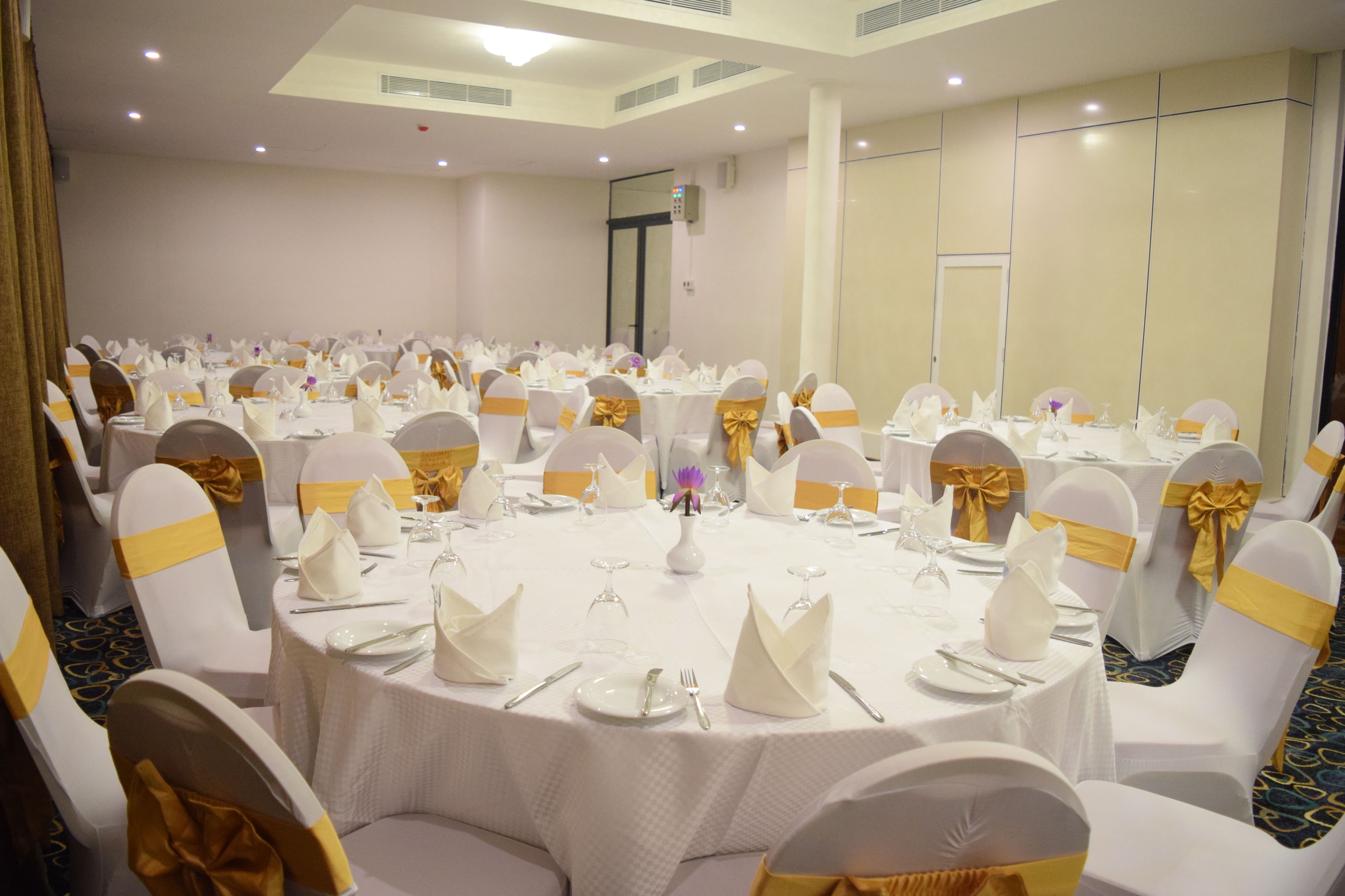 Une salle de banquet avec des tables rondes dressées pour le repas, des chaises blanches ornées de rubans dorés et une palette de couleurs neutres.